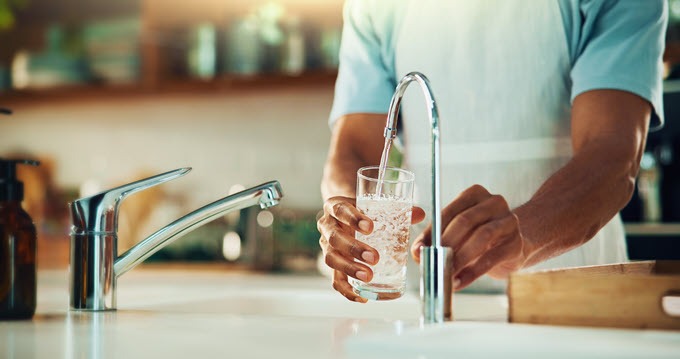 a woman stands in front of a faucet, pouring a glass of water from a water treatment system.